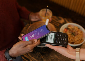 A person is using a smartphone linked to a neobank account for a contactless payment on a card reader displaying "$19.90." A bowl of noodles on the table suggests the transaction is taking place at a restaurant. | OpenBank.com